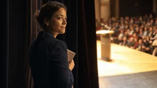 Mulher de blazer azul-marinho nos bastidores de um auditório, segurando anotações junto ao peito, olha para o palco iluminado onde a plateia espera diante do púlpito vazio