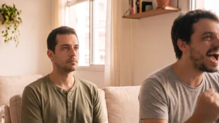 Homem sentado no sofá olha para o lado com expressão vazia enquanto o amigo, ao lado, comemora um gol de punho fechado