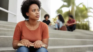 Estudante sentada na escada do campus com um livro fechado no colo olha para o lado, apreensiva, minutos antes de uma prova